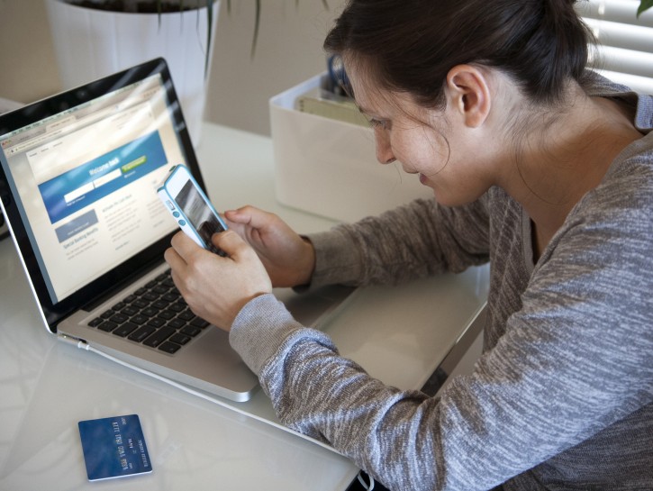 Young woman sitting at table using smart phone and laptop, credit card on table
