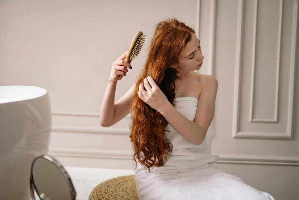 Donna che si spazzola capelli lunghi rossi mossi in sala da bagno avvolta da asciugamano bianco