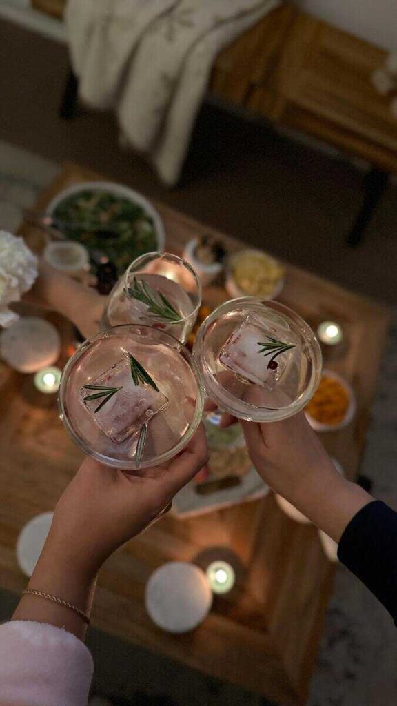 Mani di persone con cocktail con ghiaccio e rosmarino che fanno cin cin, luci soffuse