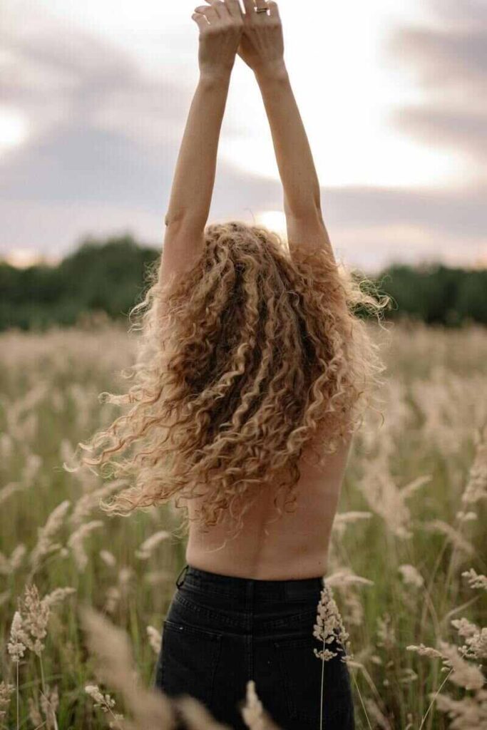 Donna di schiena in un campo con capelli biondi ricci in movimento e braccia in alto