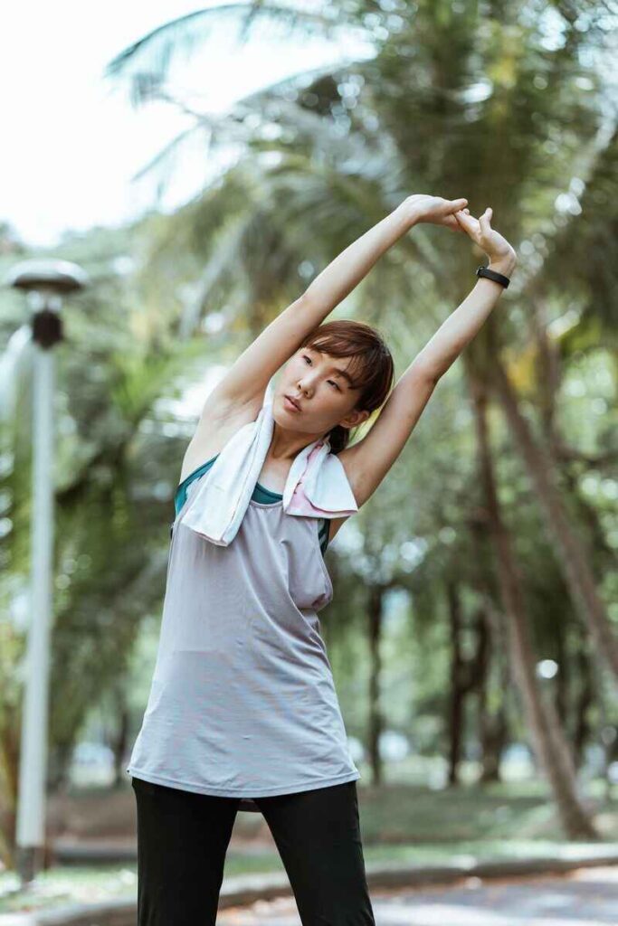 Ragazza asiatica in completo sportivo con asciugamano al collo che fa stretching allungando braccia in alto