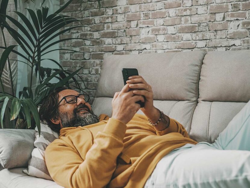 Uomo maturo sdraiato sul divano sorride guardando lo smartphone