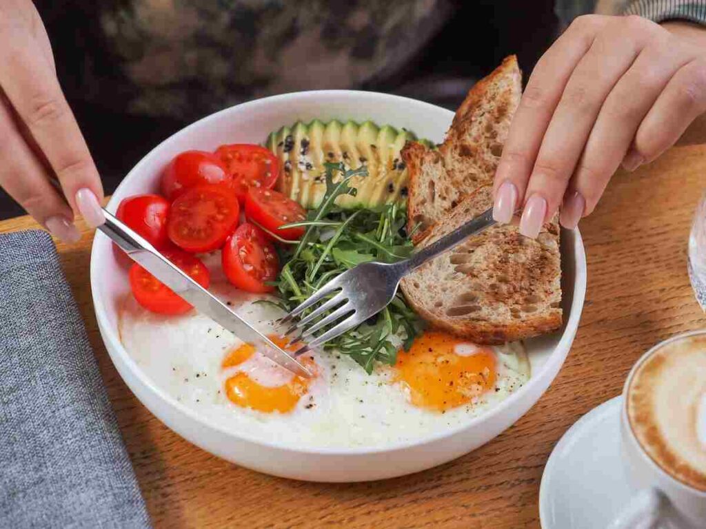 mani di donna che tagliano uova in piatto con pomodorini, avocado, rucola e pane 