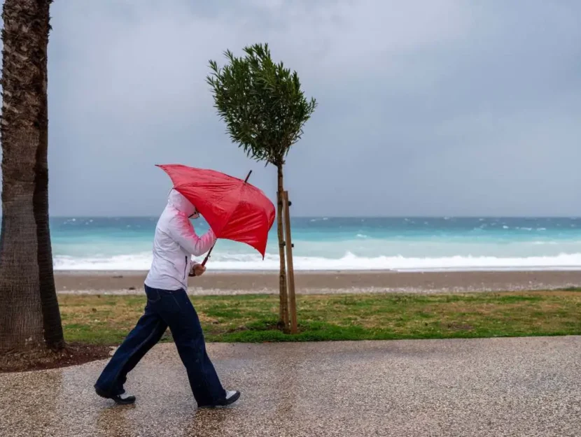 Giovane donna con un impermeabile bianco che lotta con un ombrello arcobaleno in una giornata ventosa e piovosa sul mare