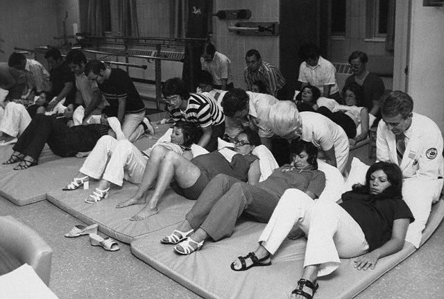 29 Dec 1971, New York State, USA --- Original caption: Family Matters. New Hyde Park, N.Y.: Fathers-to-be help, support, coach and kibbitz during pre-parenthood classes at Long Island Jewish Medical Center in New Hyde Park. The Medical Center program, before and after the child's birth, not only teaches essentials but strengthens Dad's role-and ego-through active participation in his wife's care during pregnancy and labor and through the baby's care almost from the moment it is born. In this recent exercise class at the medical center, fathers give physical and emotional support to their wives. This exercise is designed to help in last stages of delivery. Helping at extreme right is Dr. Fred Benjamin of Great Neck, director of Obstetrics/Gynecology and RN Ruth Martins, assistant director of nursing for Obstetrics/Gynecology. --- Image by © Bettmann/CORBIS